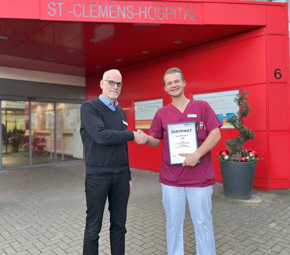 Ludger Broeckmann, Kaufmännischer Direktor des St.-Clemens-Hospitals Geldern, gratuliert Tim Möller, examinierte Pflegefachkraft im St.-Clemens-Hospital, mit einem Händedruck vor dem Eingang des Krankenhauses zu seiner erfolgreich bestandenen Weiterbildung als Social Media Nurse. Tim Möller hält dabei das Zertifikat der Weiterbildung in der Hand.